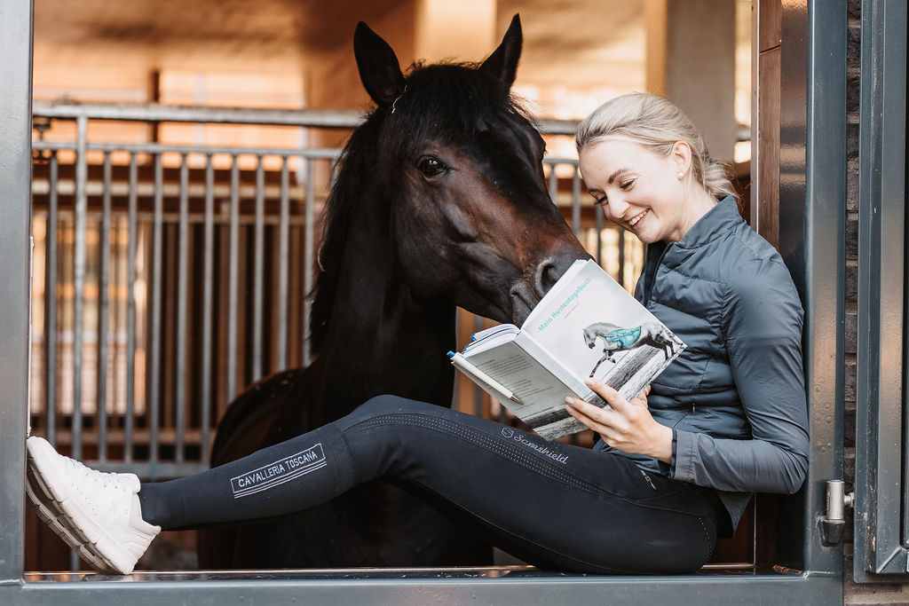 Frau sitzt auf Stallfenster und schaut mit Pferd in Hustentagbuch