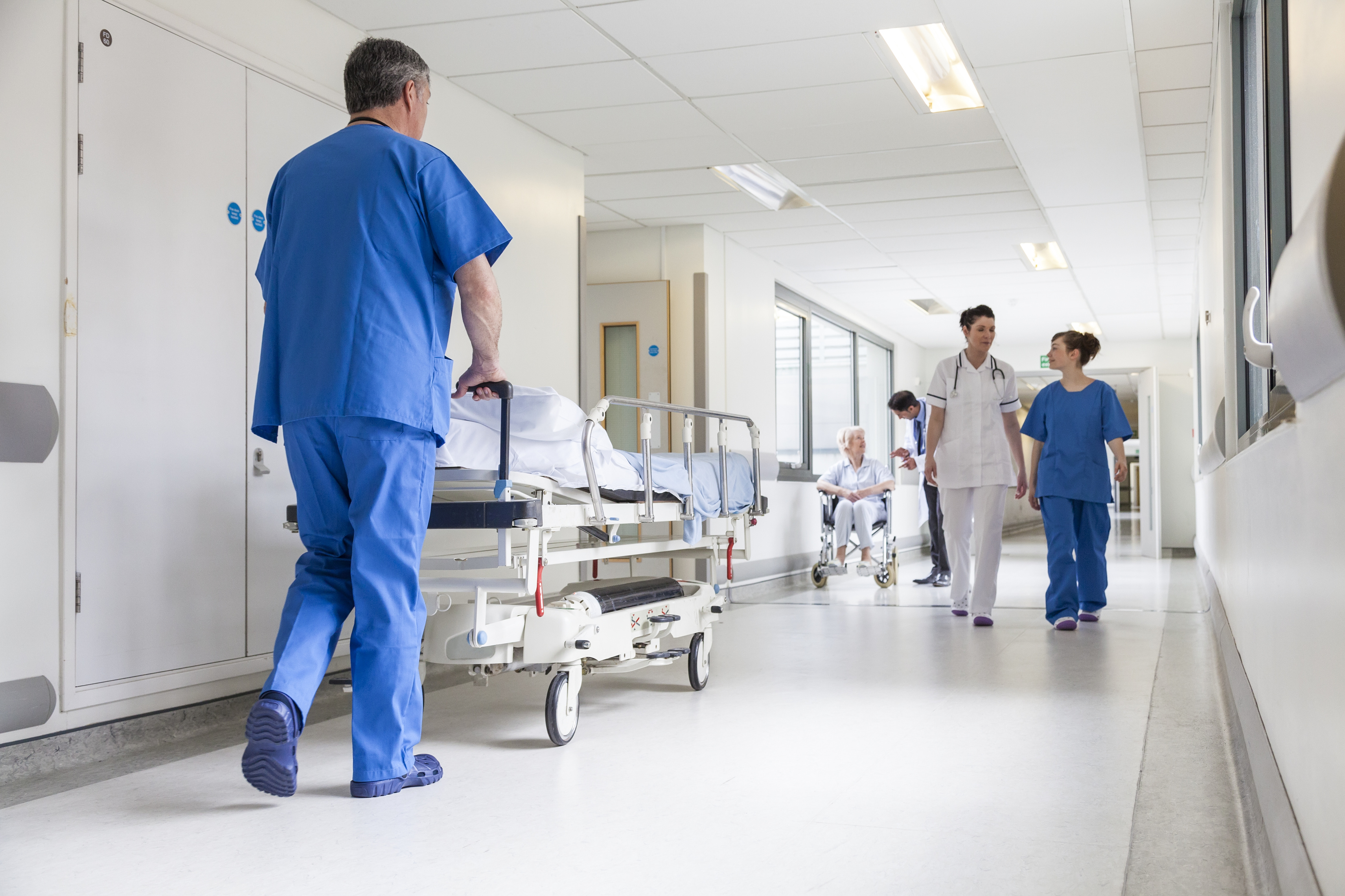 © 54048895 - spotmatikphoto - stock.adobe.com Krankenpfleger schiebt Krankenhausbett im Flur, während Kollegen vorbei gehen